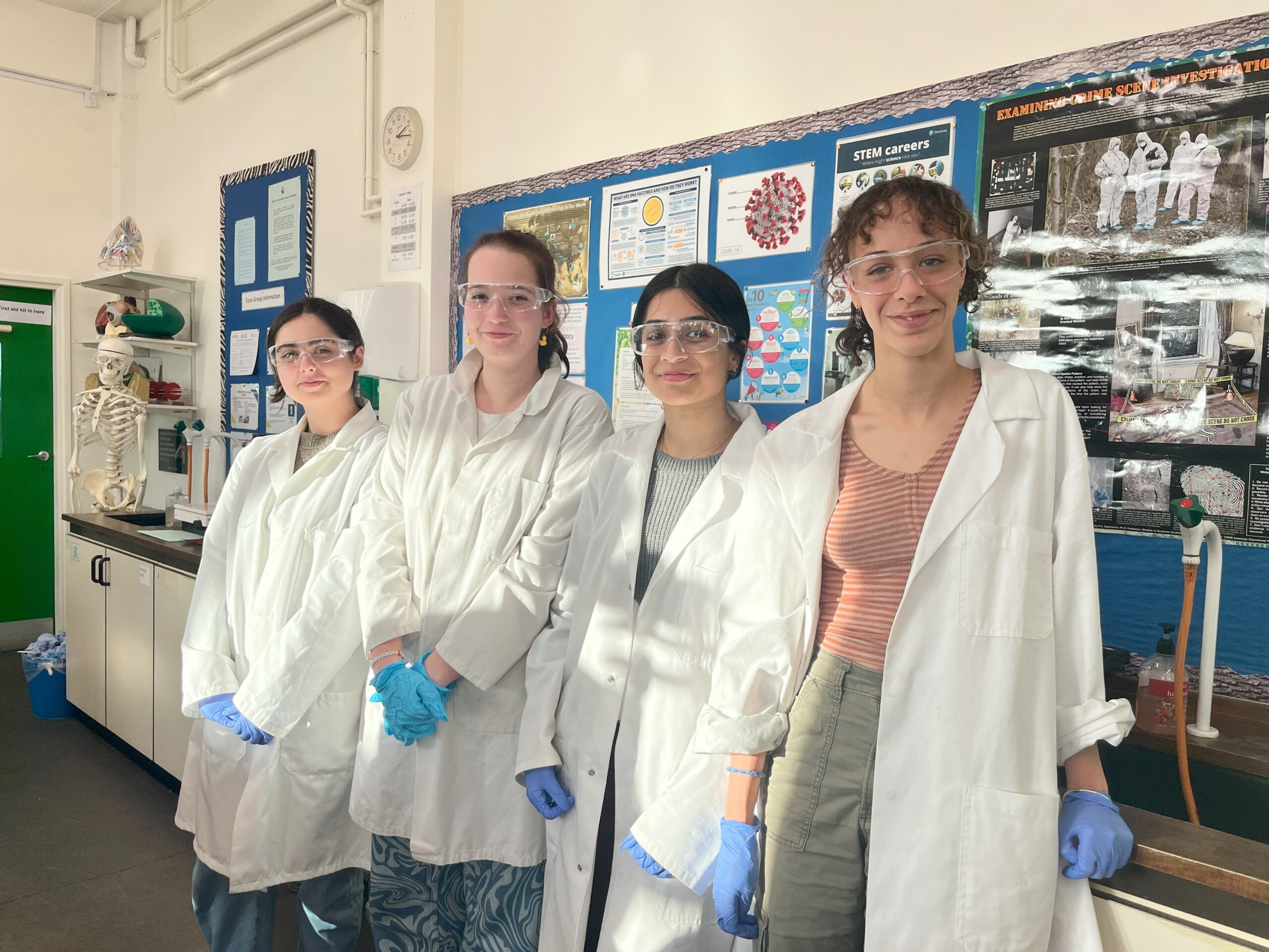 Four female Biology students, wearing googles and in white lab coats smiling