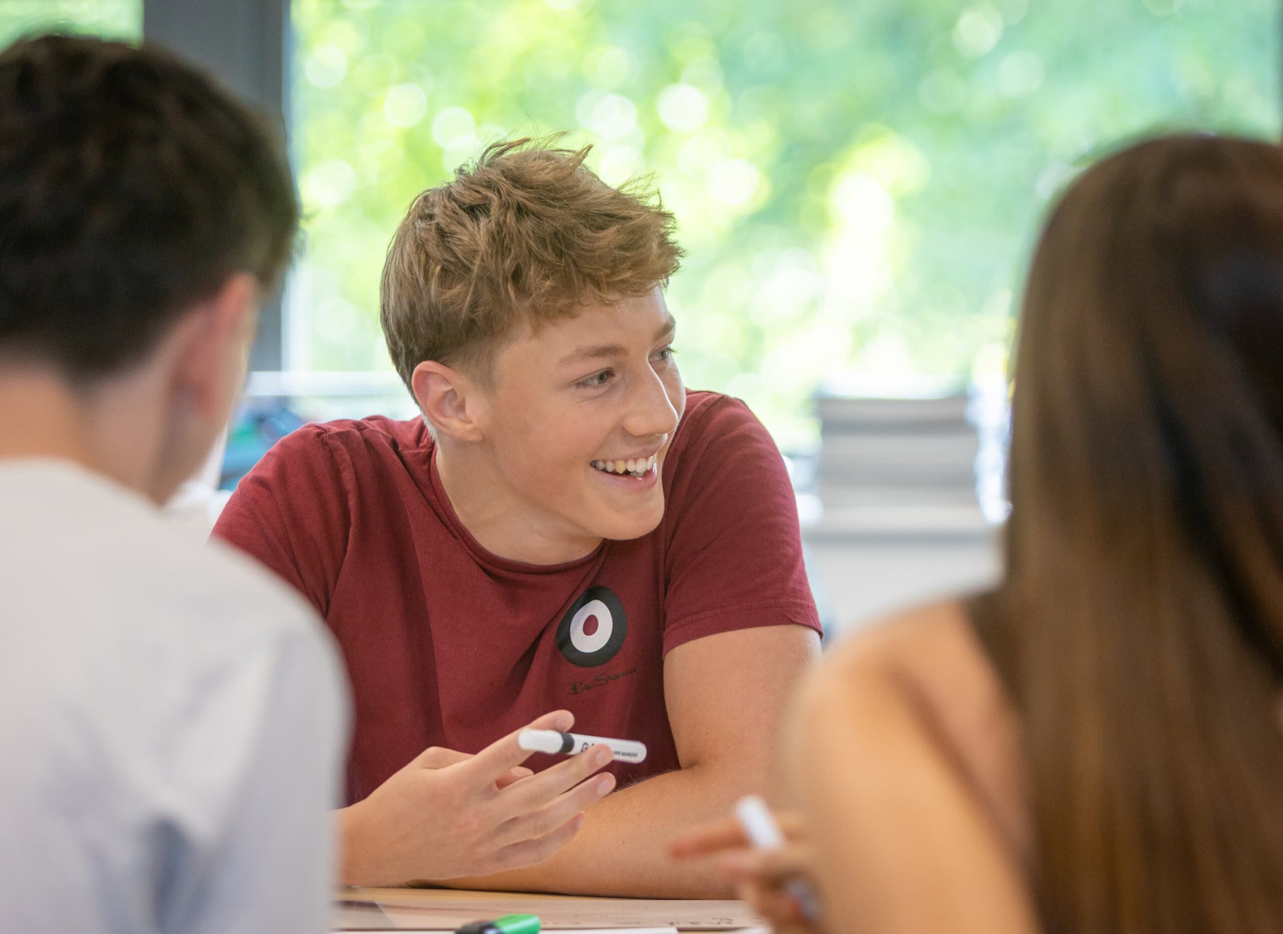 Boy sat at a table in a classroom studying and talking with friends