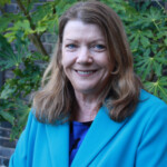 Photo of a woman standing by a bush, wearing a blue coat and smiling