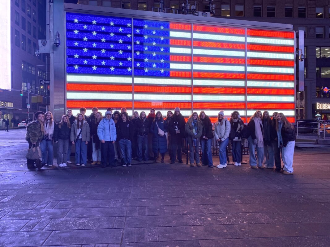 Students stand infront of a digital bilboard showing the American Flag in New York City's Times quare