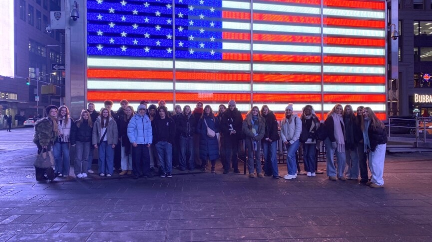 Students stand infront of a digital bilboard showing the American Flag in New York City's Times quare