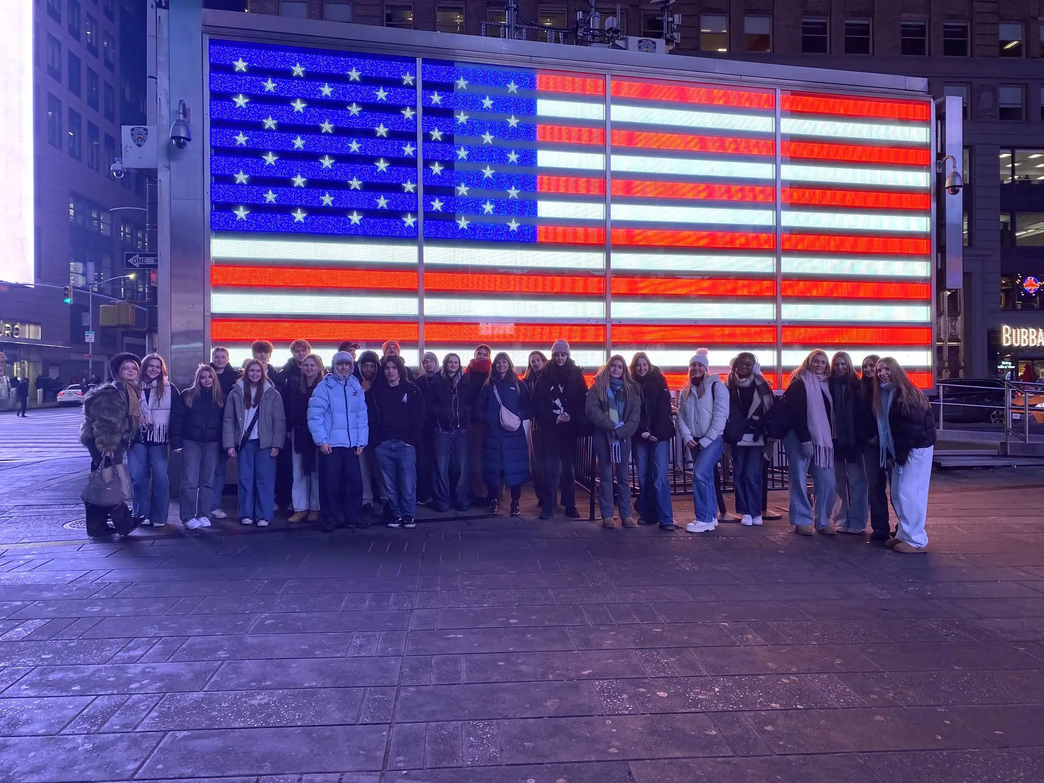 Students stand infront of a digital bilboard showing the American Flag in New York City's Times quare