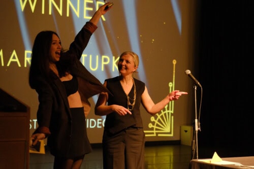 An excited student holding a trophy standing next to a female teacher on stage