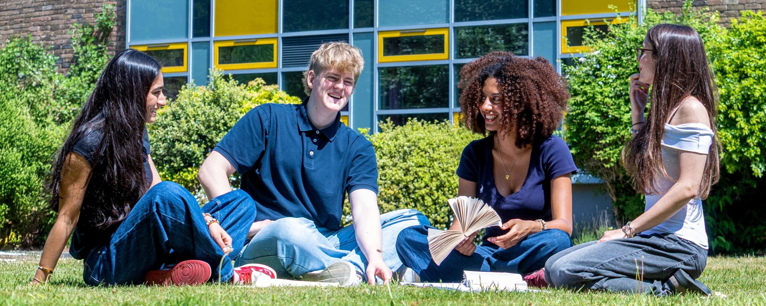 three girls and a boy sit on the grass talking with their books outside esher sixth form college