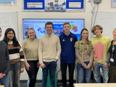 A group of geography students, teachers and guest speakers stand in a line in a classroom