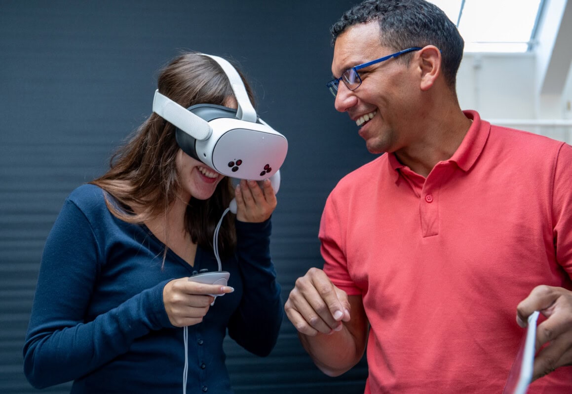 male teacher in pink shirt, smiles with a female graphic communications student as she tries on a VR headset