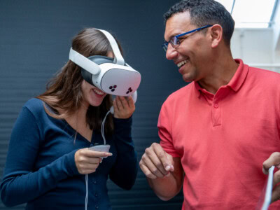 male teacher in pink shirt, smiles with a female graphic communications student as she tries on a VR headset