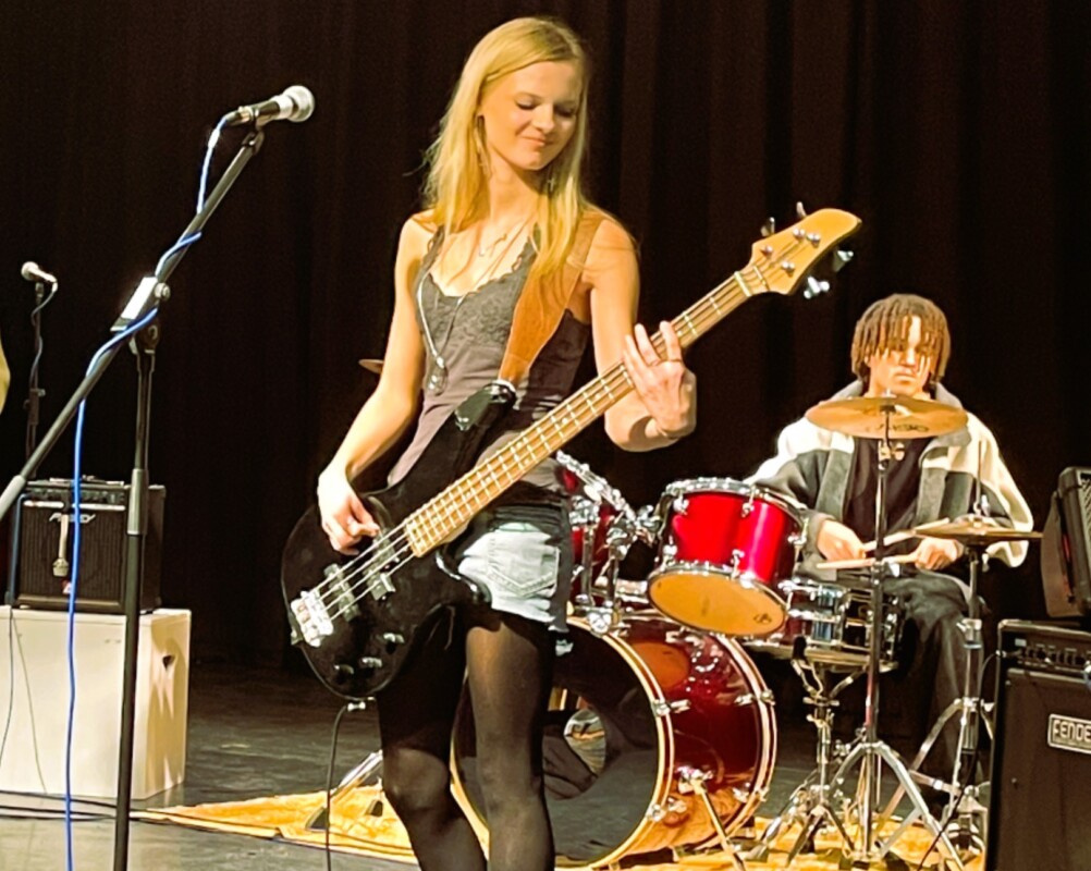 Girl plays Guitar with boy playing drums behind for College Band