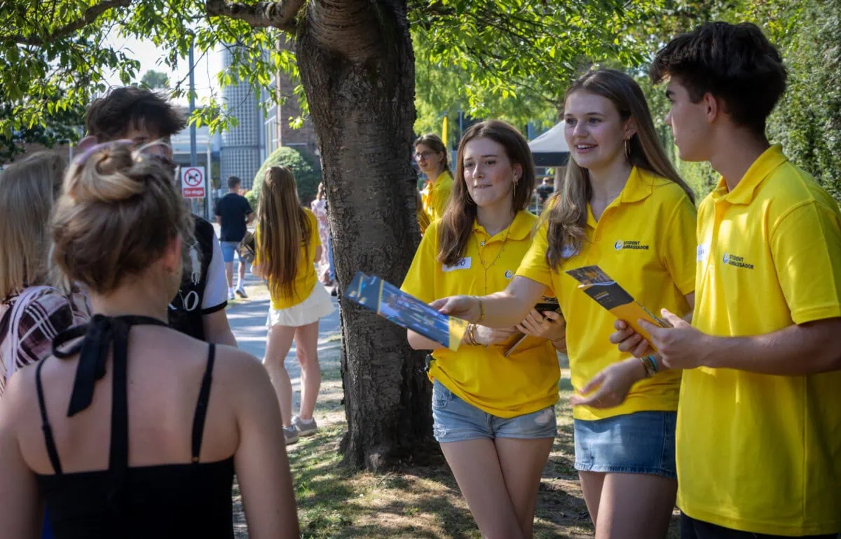three students wearing yellow polo shirts hand out prospectuses to visitors