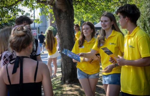 three students wearing yellow polo shirts hand out prospectuses to visitors